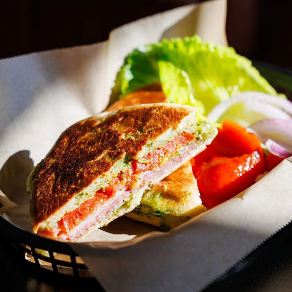 Grilled sandwich layered with tomato, cheese, and greens, served with lettuce, onions, tomato, and pickles in a lined basket.